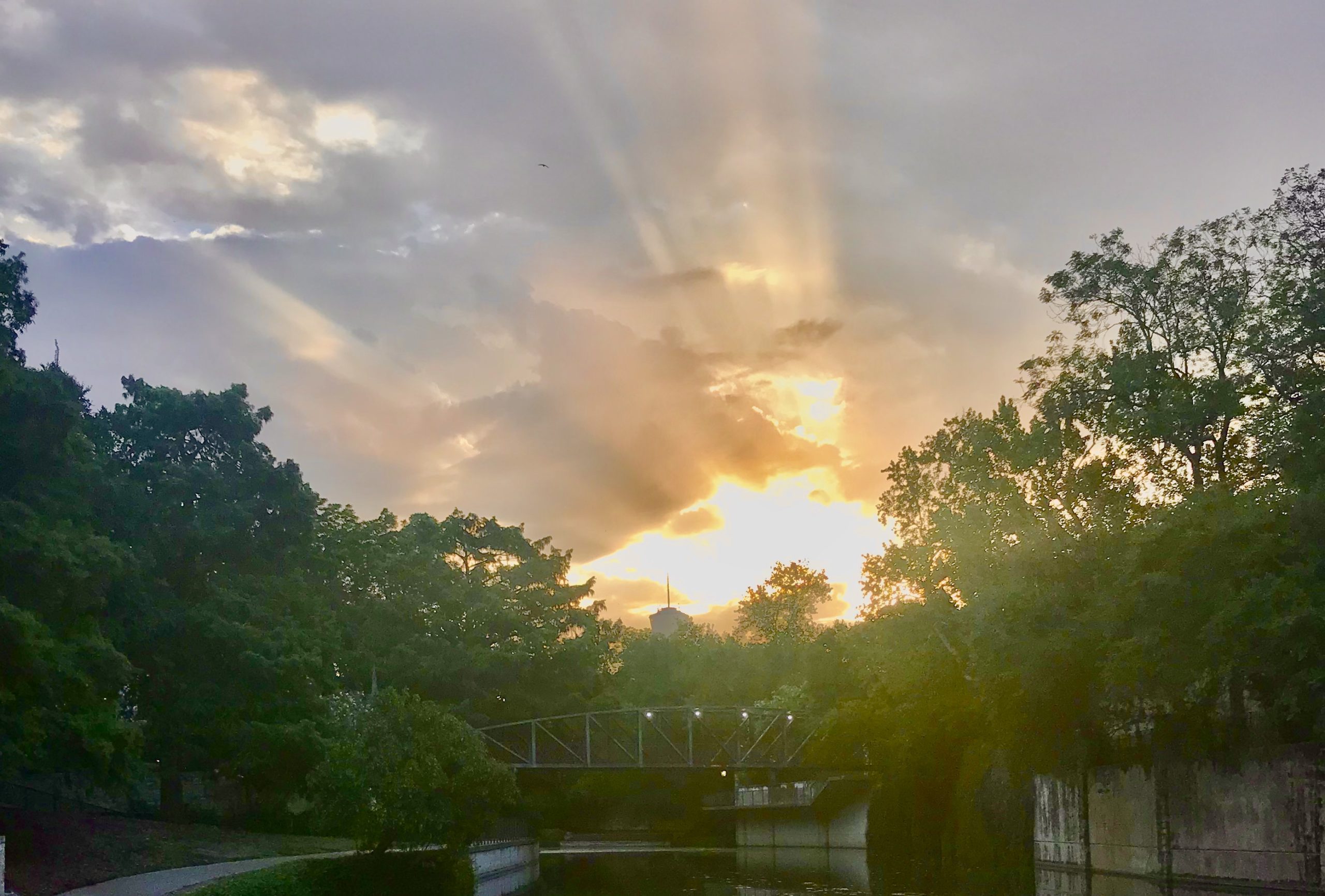 hero image of sunset bursting with rays over a green treeline, with the san antonio river in foreground.