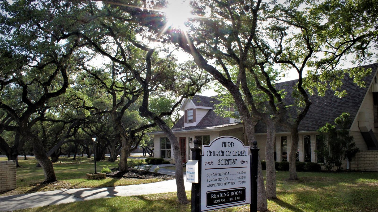 photo of the front of Third Church Christ Scientist, San Antonio. In foreground: A welcoming sign with the text "Third Church of Christ Scientist" with hours. Many beautiful oak trees shade the building, and sun beams shine through the trees at top center.