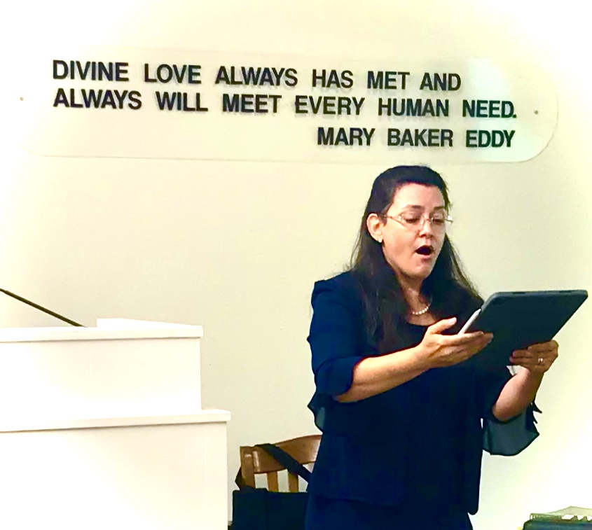 K. Stephenson holding a black binder open and singing during a church service in front of a white wall, under the words "Divine Love Always Has Met And Always Will Meet Every Human Need. Mary Baker Eddy"